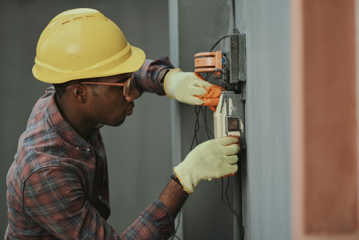 An electrician in a hard hat and gloves wiring a wall panel with both hands